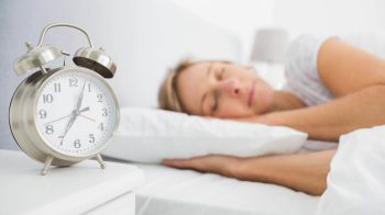 Blonde woman asleep in bed while her alarm shows the early time at home in bedroom 2 e1653865363692 How Many Hours Of Sleep Should You Be Getting?